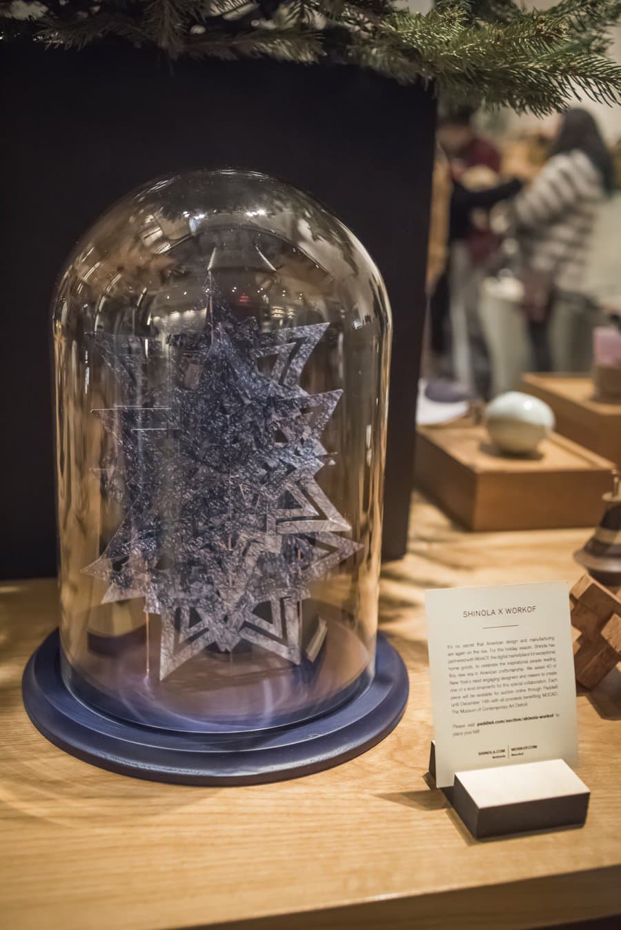 Cut paper snowflake ornament under a glass dome on a wood display at the Shinola tree lighting