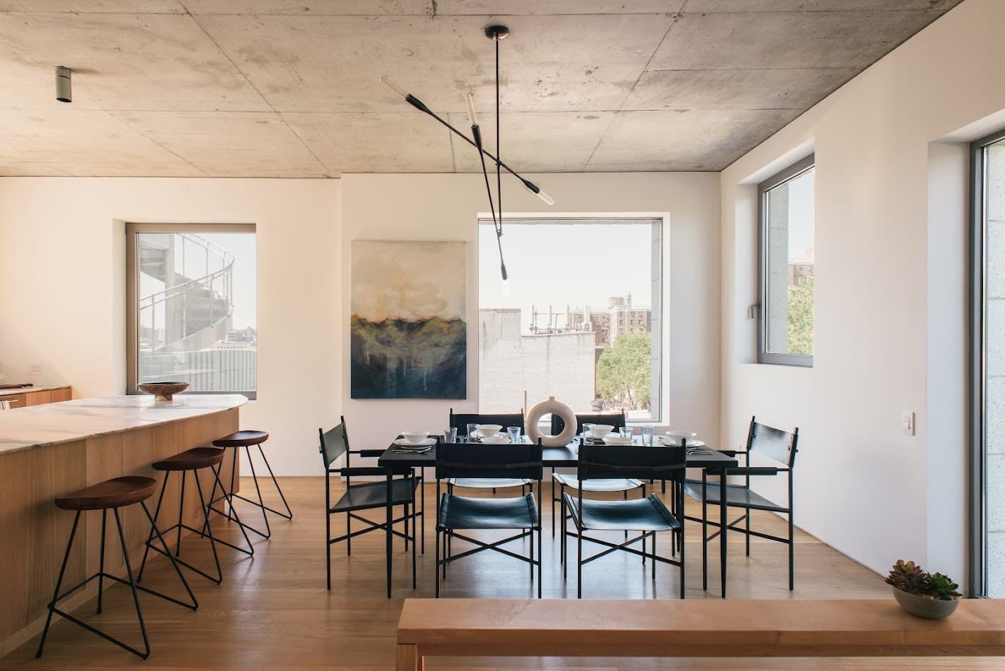 Dining room in the 450 Warren penthouse in Gowanus, Brooklyn, with black leather chairs, a sculptural pendant, an abstract painting and an oak kitchen island under an exposed concrete ceiling