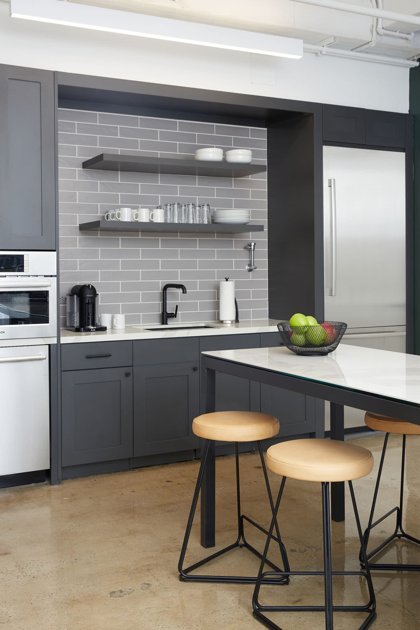 Office kitchen with grey cabinetry, open shelves on grey tile, and leather-seated stools at a white island