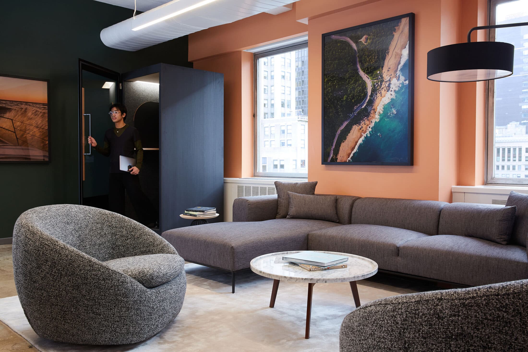 BRE office lounge with terracotta walls, a charcoal sectional, a bouclé swivel chair, and a large aerial beach photograph