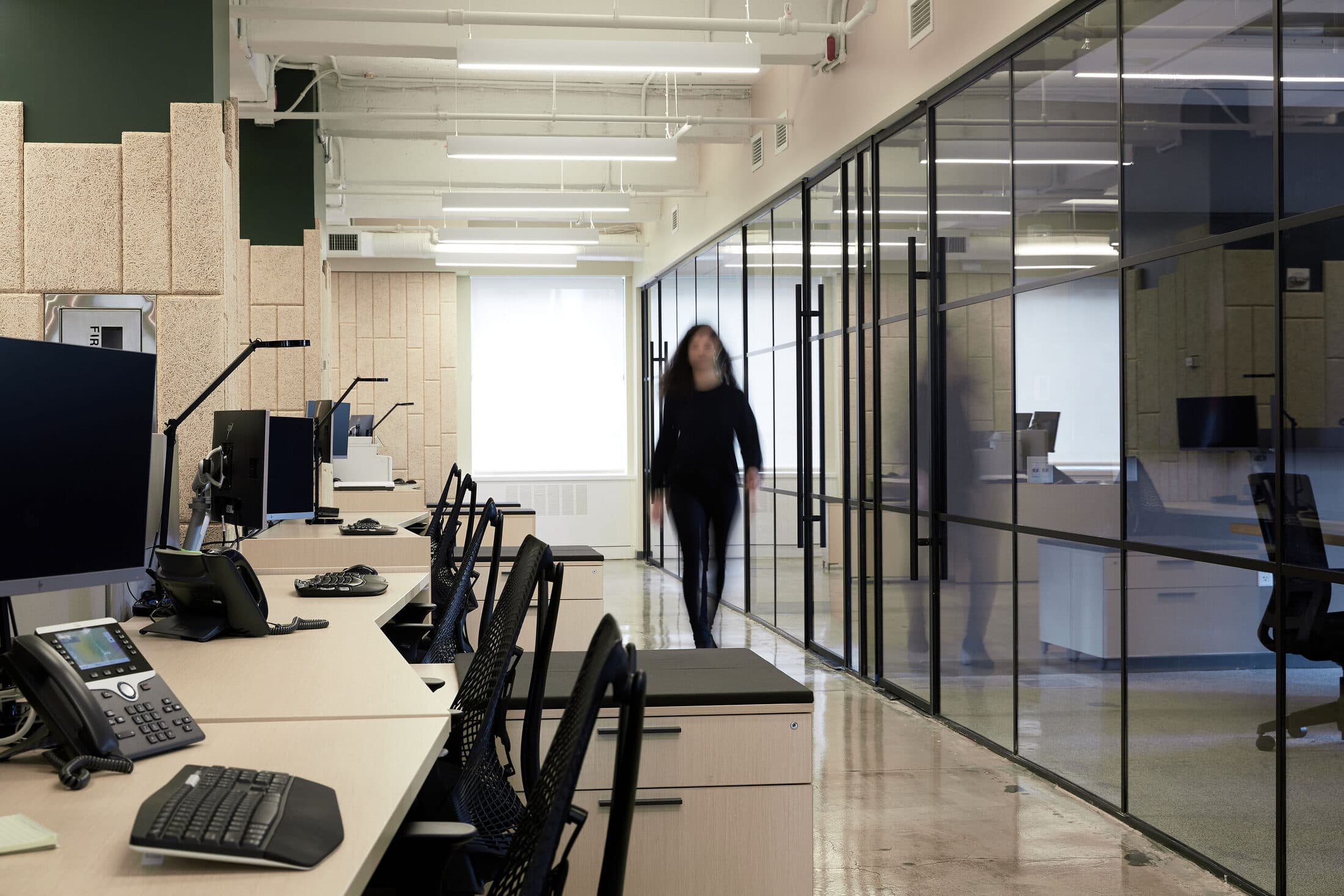 Open office with long white desks, black task chairs, and glass-walled offices along one side
