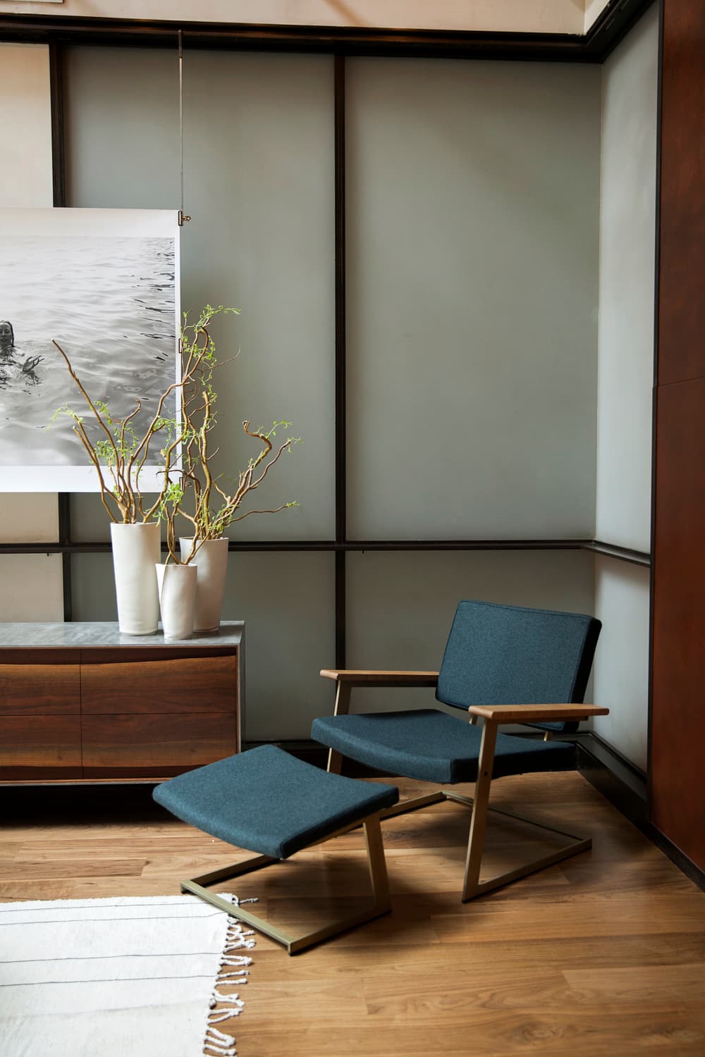 Slate-blue lounge chair and ottoman beside a walnut credenza with white ceramic vessels and curly willow branches against grey painted panels