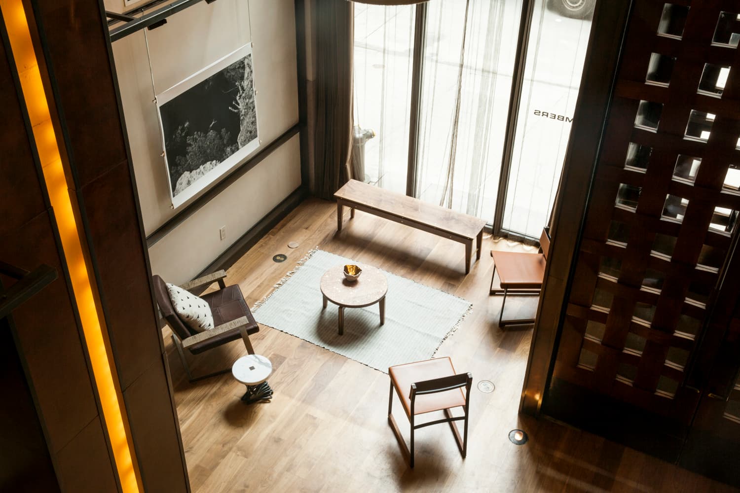 View from the mezzanine down to a Chambers Hotel lounge grouping by the tall street windows, with a bench, a round side table, and leather chairs on a white rug