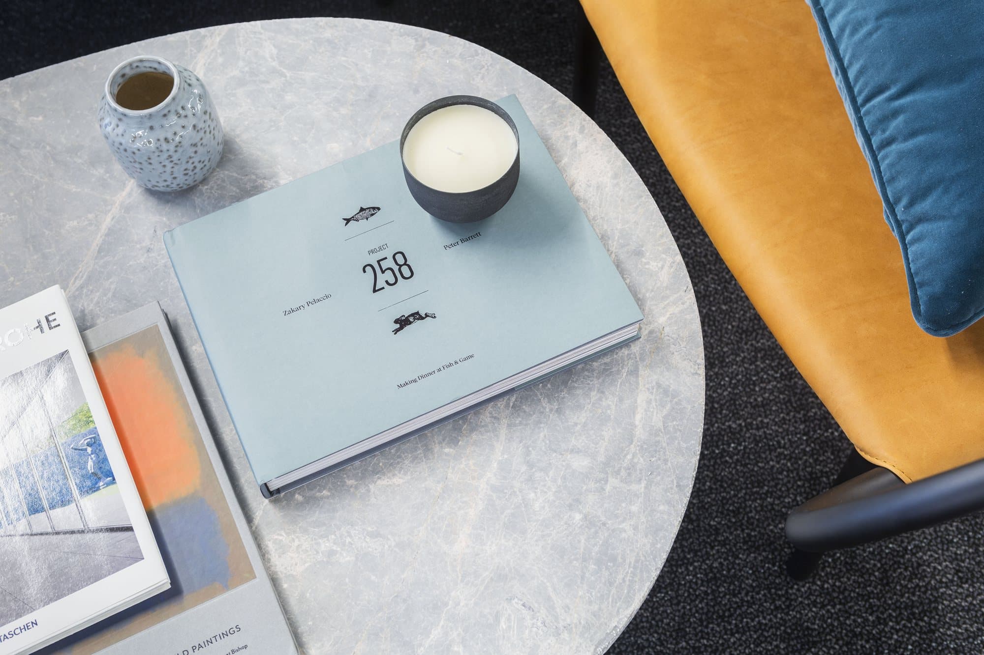 Close-up of coffee table books, a ceramic cup and a candle on a marble table beside cognac and teal upholstery