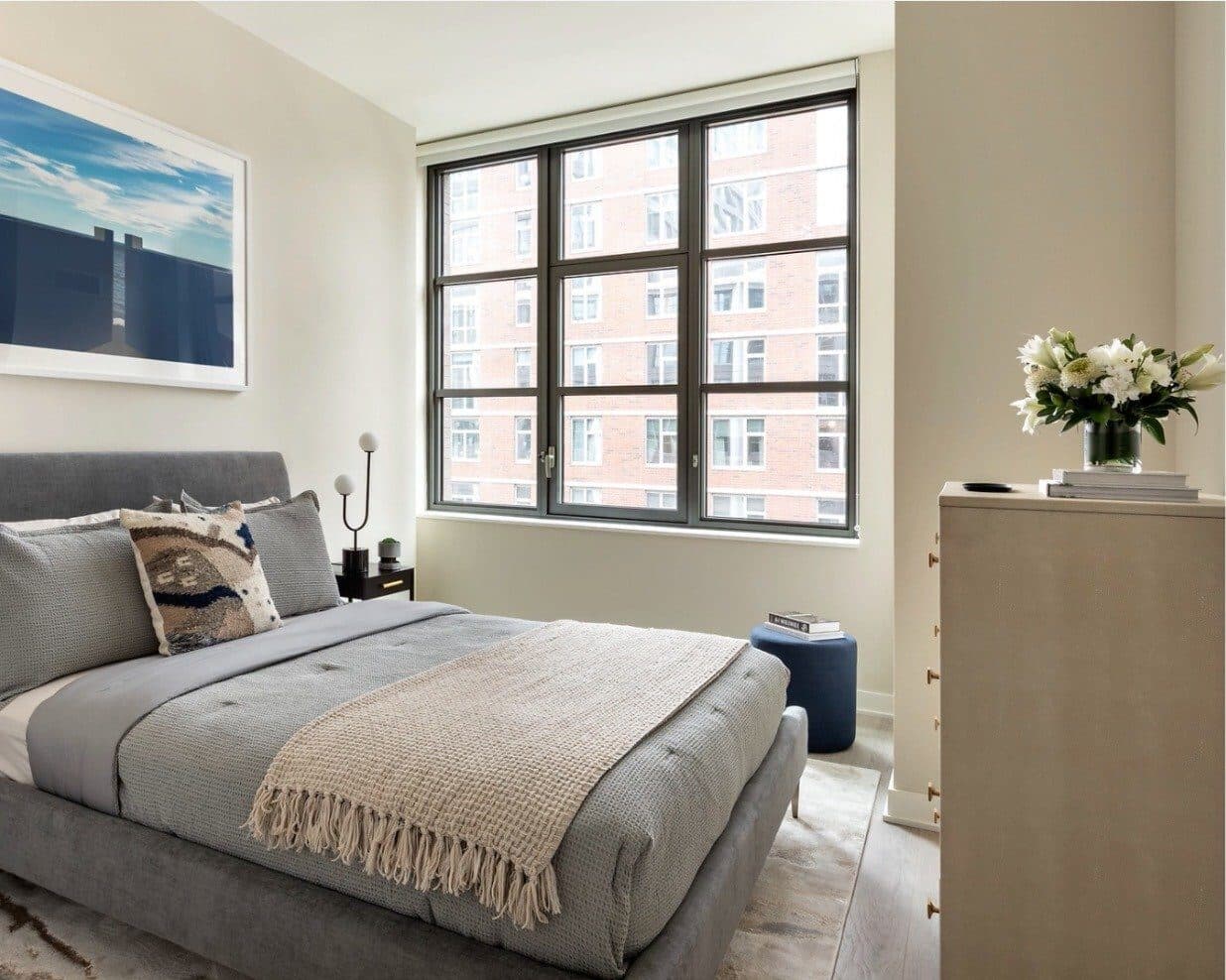 Bedroom with a grey upholstered bed, a blush throw and a tall dresser by the window