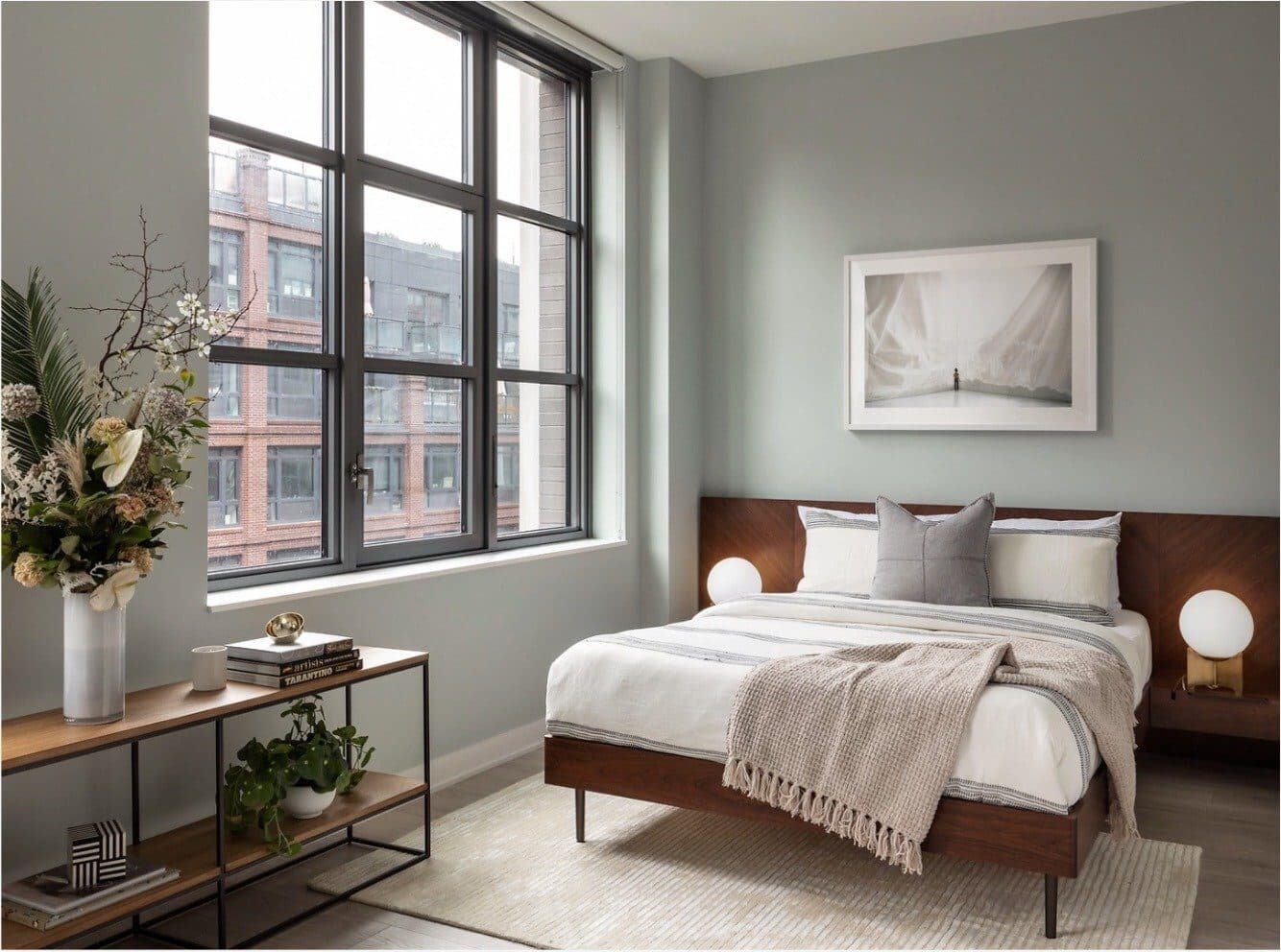 Studio bed with a walnut frame and cream linens beside a console with plants and a framed photograph