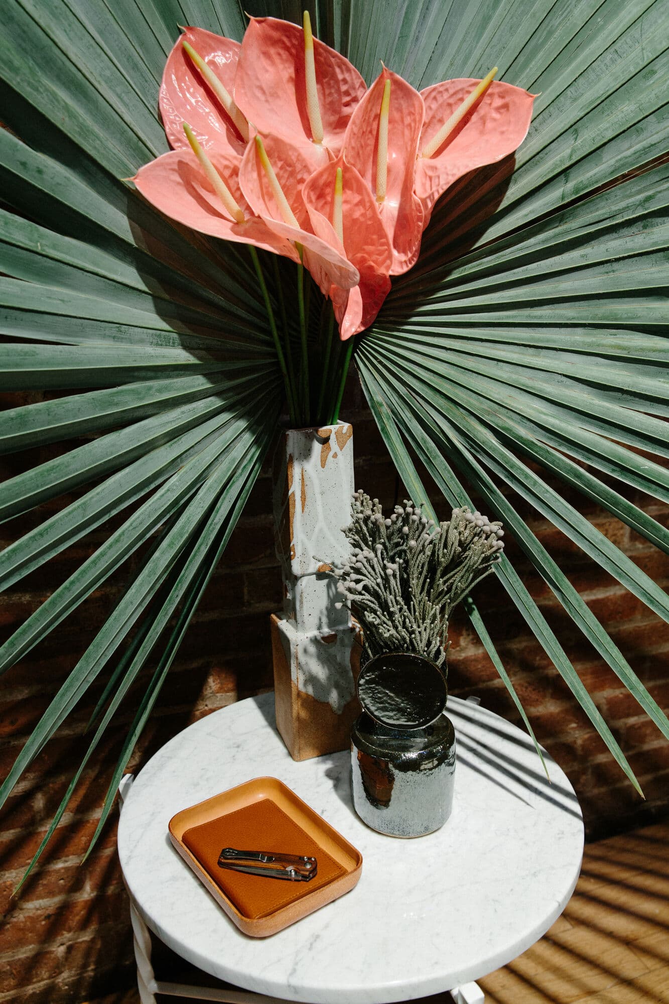 Coral anthurium against a fan palm leaf in a stone vase, beside a Shinola leather tray on a white marble side table