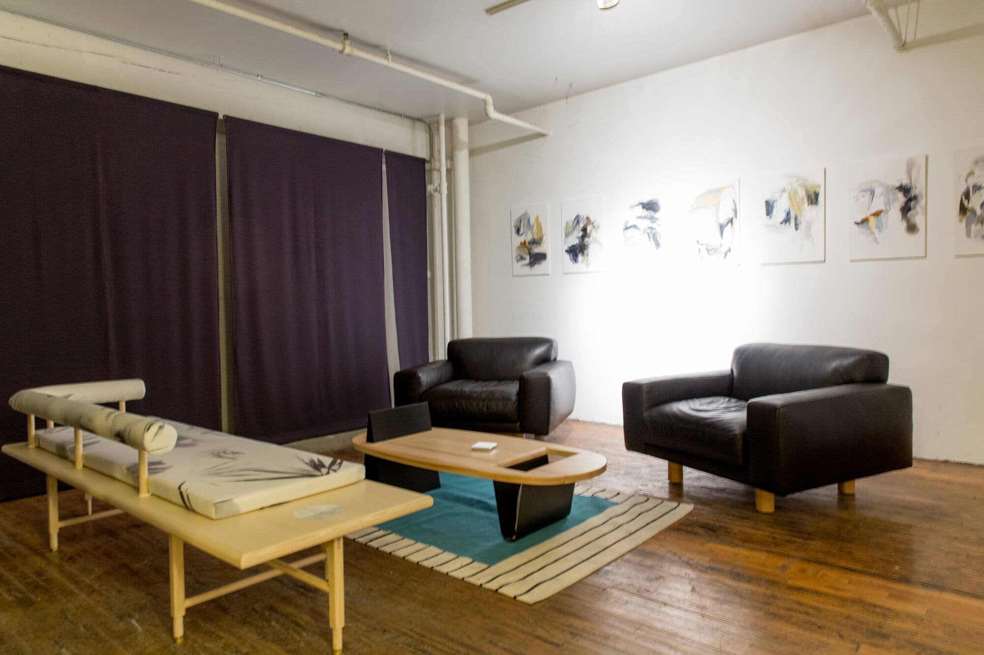Lounge with black leather armchairs, a light oak bench and a low table on a teal rug beneath a row of botanical prints