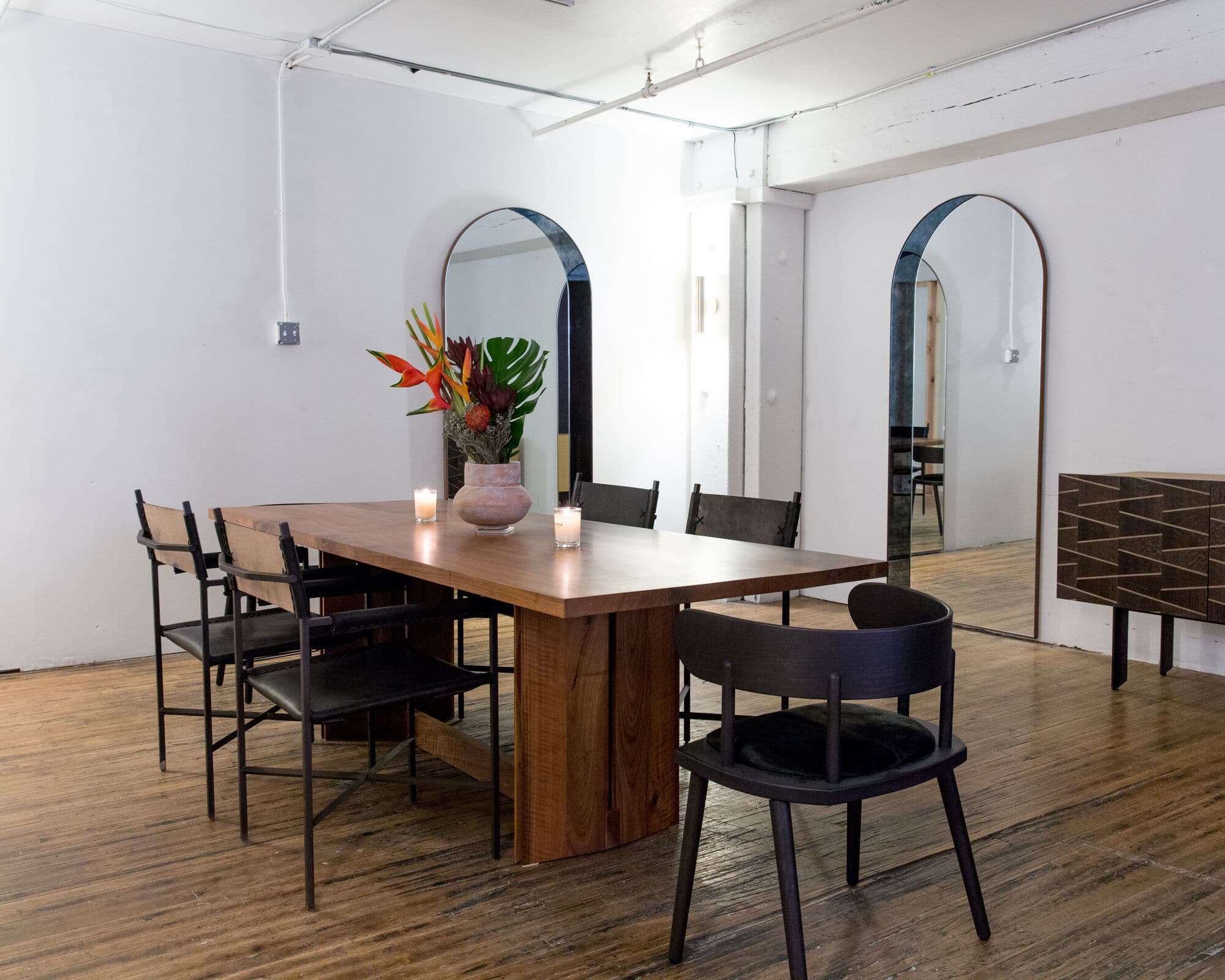 Walnut dining table with black chairs, candles and tropical flowers in a loft with arched mirrors and a marquetry credenza